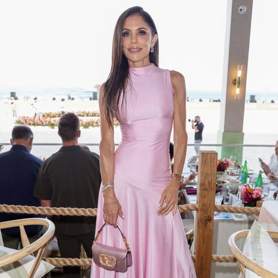 A woman in a sleeveless pink dress stands indoors by a table, holding a small pink purse. People are seated behind her, and windows reveal a bright outdoor view.