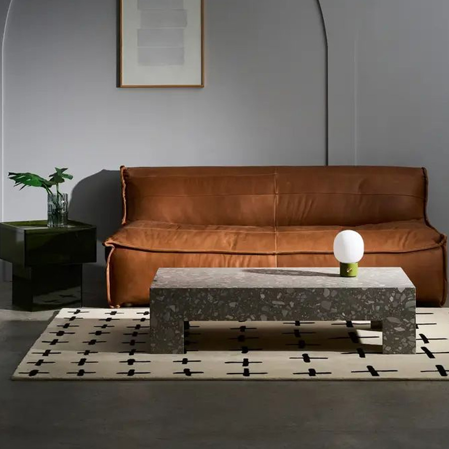 A modern living room with a brown leather sofa, terrazzo coffee table, round lamp, dark side table with a leafy plant, geometric rug, and minimalist wall art against a gray background.