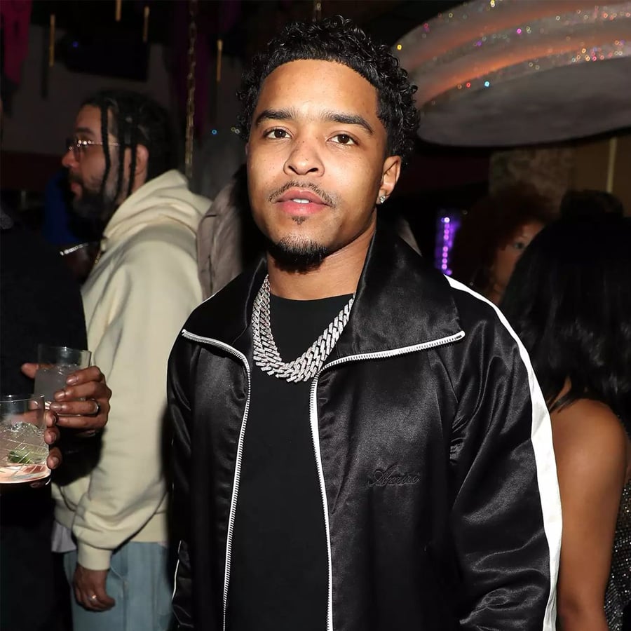 A man wearing a black tracksuit and silver chain stands in a dimly lit room at a social event. People are in the background, some holding drinks, with soft lighting and party decor visible.
