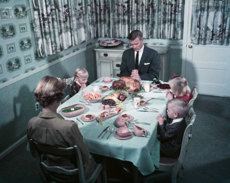 A family of five sits around a table set with food, bowing their heads in prayer before a meal in a vintage, pastel-colored dining room.