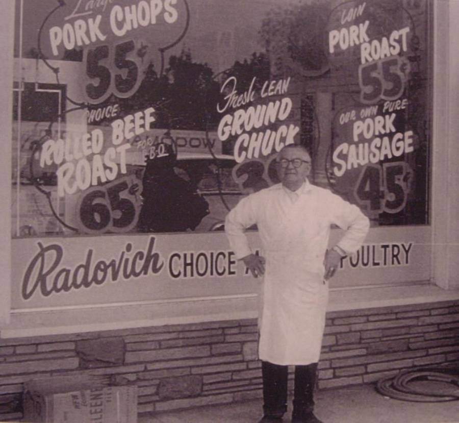 A butcher in a white apron stands outside a butcher shop with large signs advertising pork chops, beef roast, ground chuck, pork roast, and sausage prices in the window behind him.