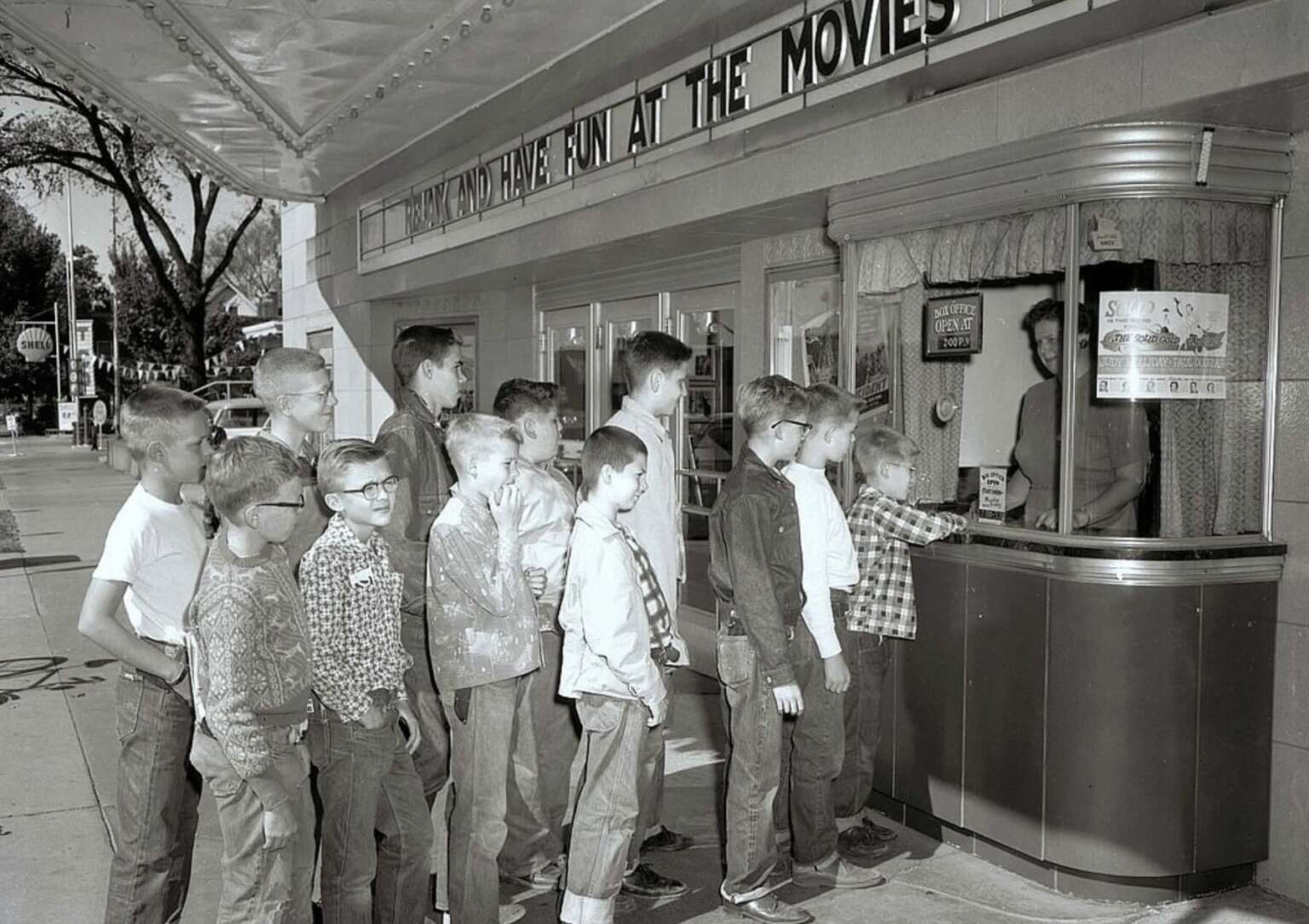 A group of boys in 1950s attire wait in line at a movie theater ticket booth, with a cashier behind the glass. The theater marquee above reads, "AND HAVE FUN AT THE MOVIES.