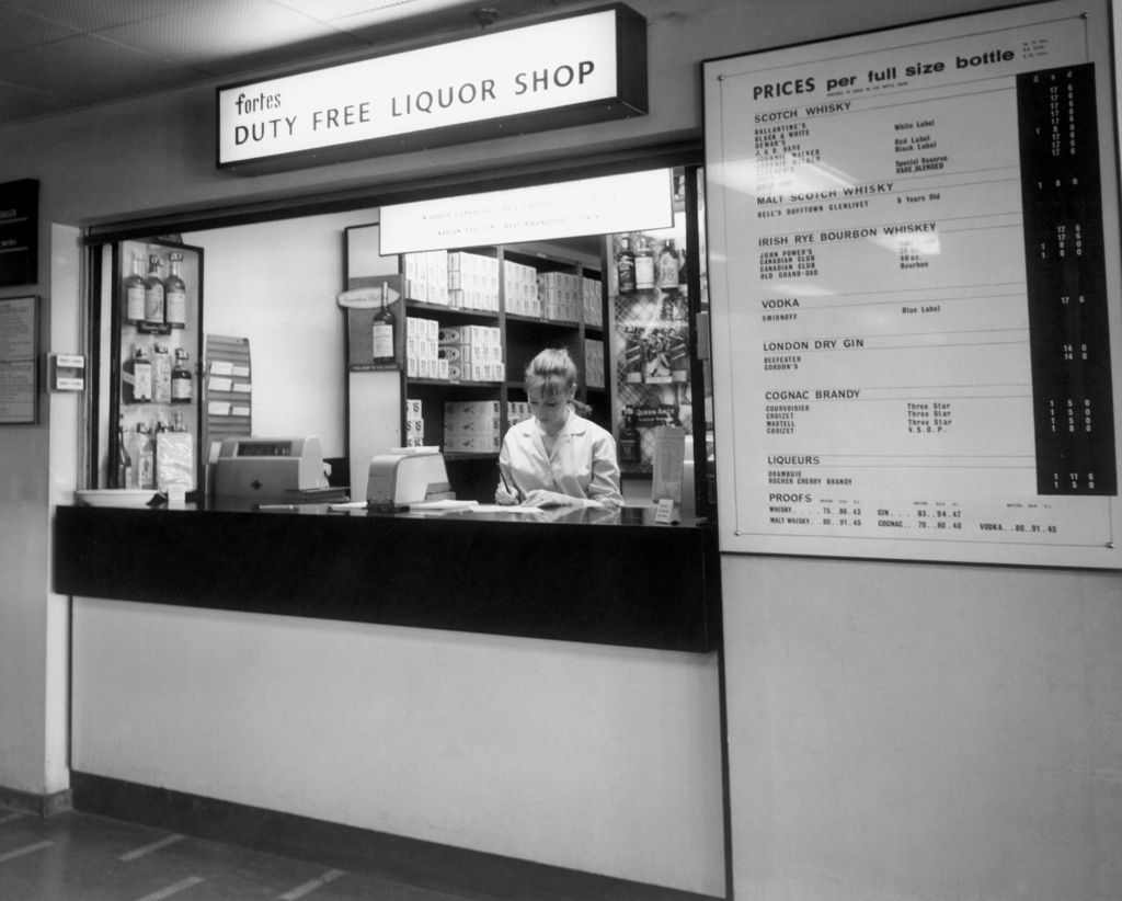 A woman stands behind the counter of a duty free liquor shop, surrounded by shelves of bottles and boxes. A large sign displays prices for various alcoholic beverages on the wall to her right.