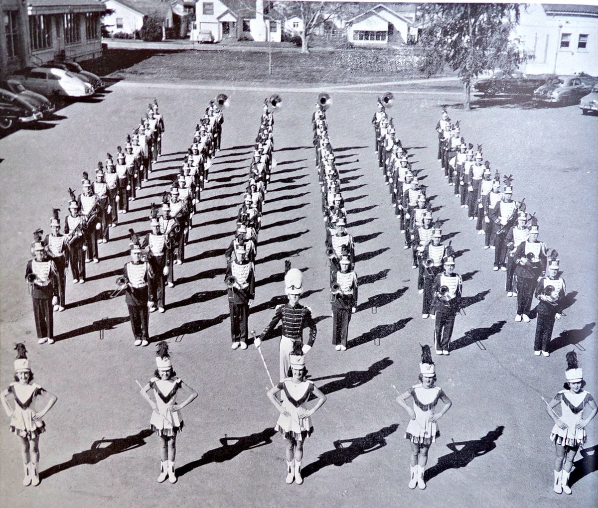A black and white photo shows a marching band in uniform, arranged in rows with baton twirlers at the front, standing in a parking lot with buildings and cars in the background.