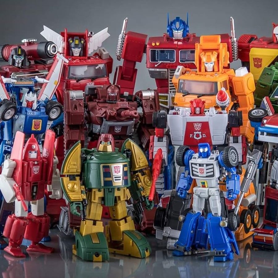 A group of colorful Transformers action figures in robot forms stands closely together on a reflective surface against a gray background.