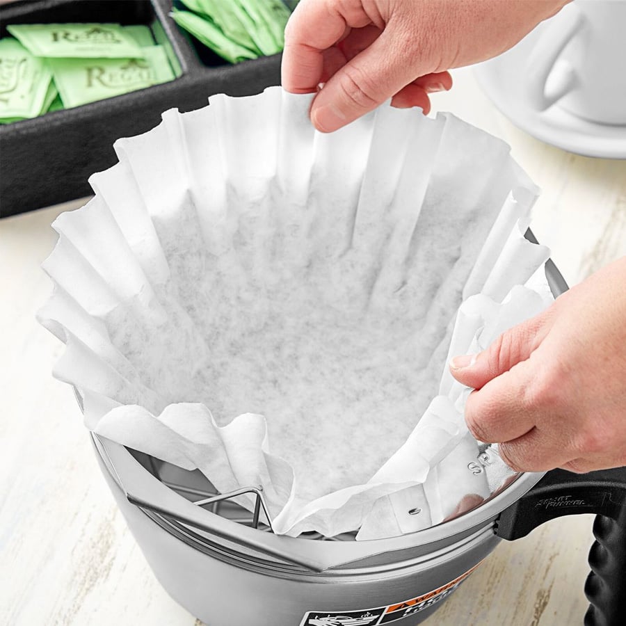 A person places a white paper coffee filter into the basket of a coffee maker, with sugar packets visible in the background.