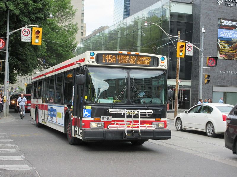 A Toronto Transit Commission 145A Humber Bay Express bus waits at a city intersection. A cyclist rides beside it, and cars and pedestrians are nearby. Tall buildings and traffic signals are visible in the background.