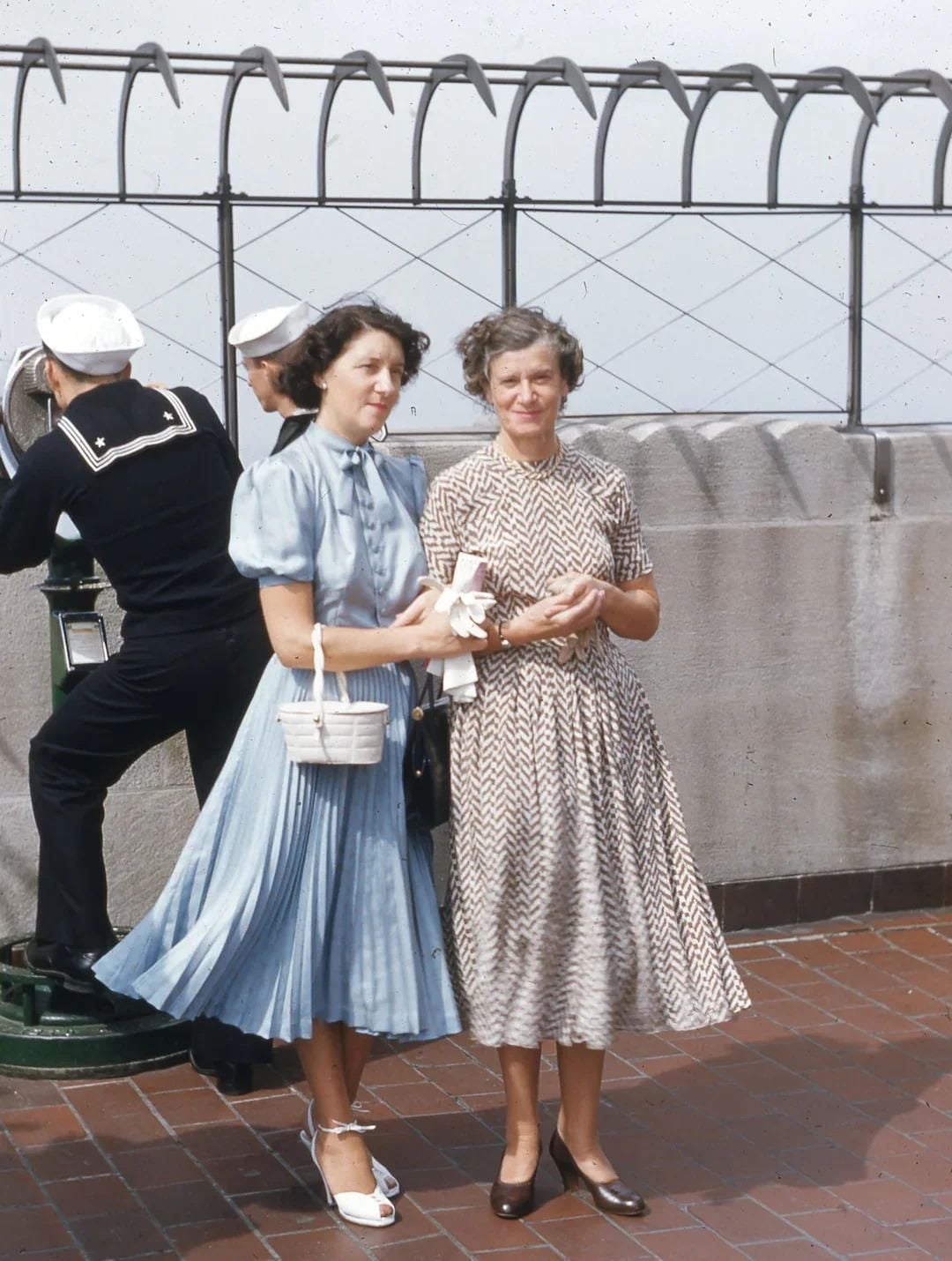 Two women in vintage dresses pose on an observation deck. One wears a blue dress with white accessories, the other a patterned dress. A man in sailor attire uses a viewing machine in the background. The scene is outdoors.