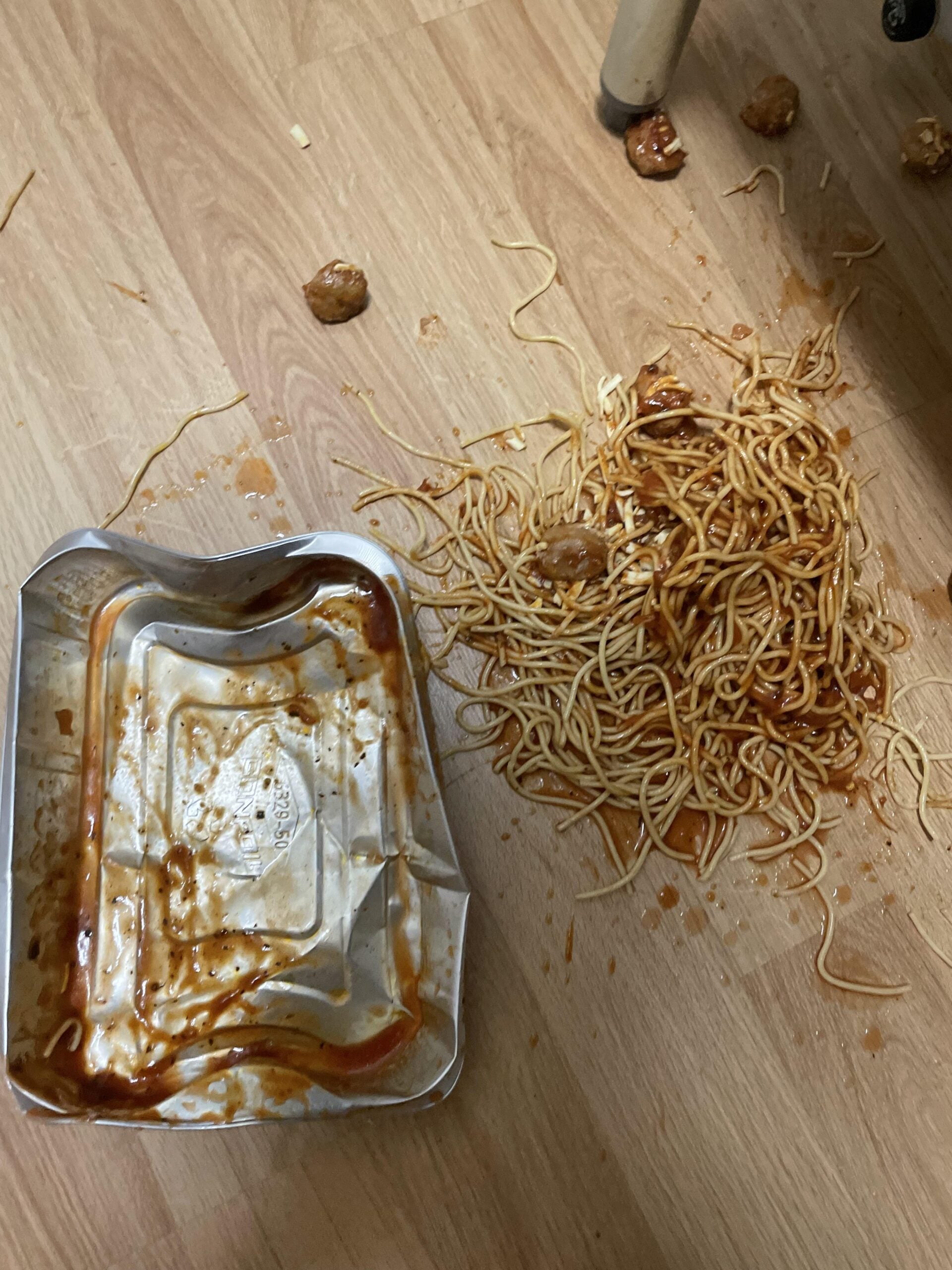 Spilled noodles and meatballs with sauce on a wooden floor next to an empty, tipped-over food container.