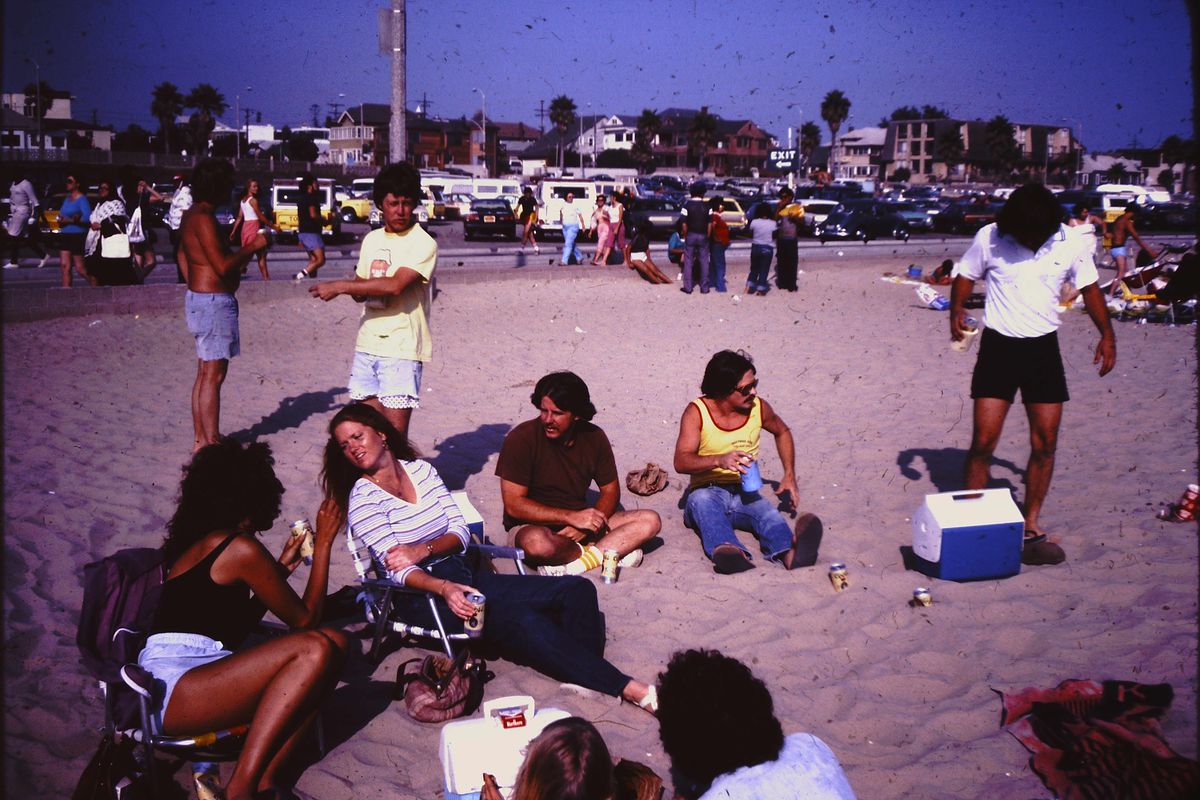 A group of people relax and socialize on a sandy beach with coolers and drinks, while others walk and sit nearby. Parked cars and houses line the background under a sunny sky.