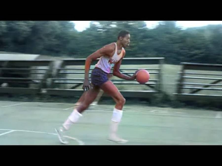 A basketball player in a white jersey and shorts dribbles a basketball on an outdoor court, with wooden bleachers and trees in the background.