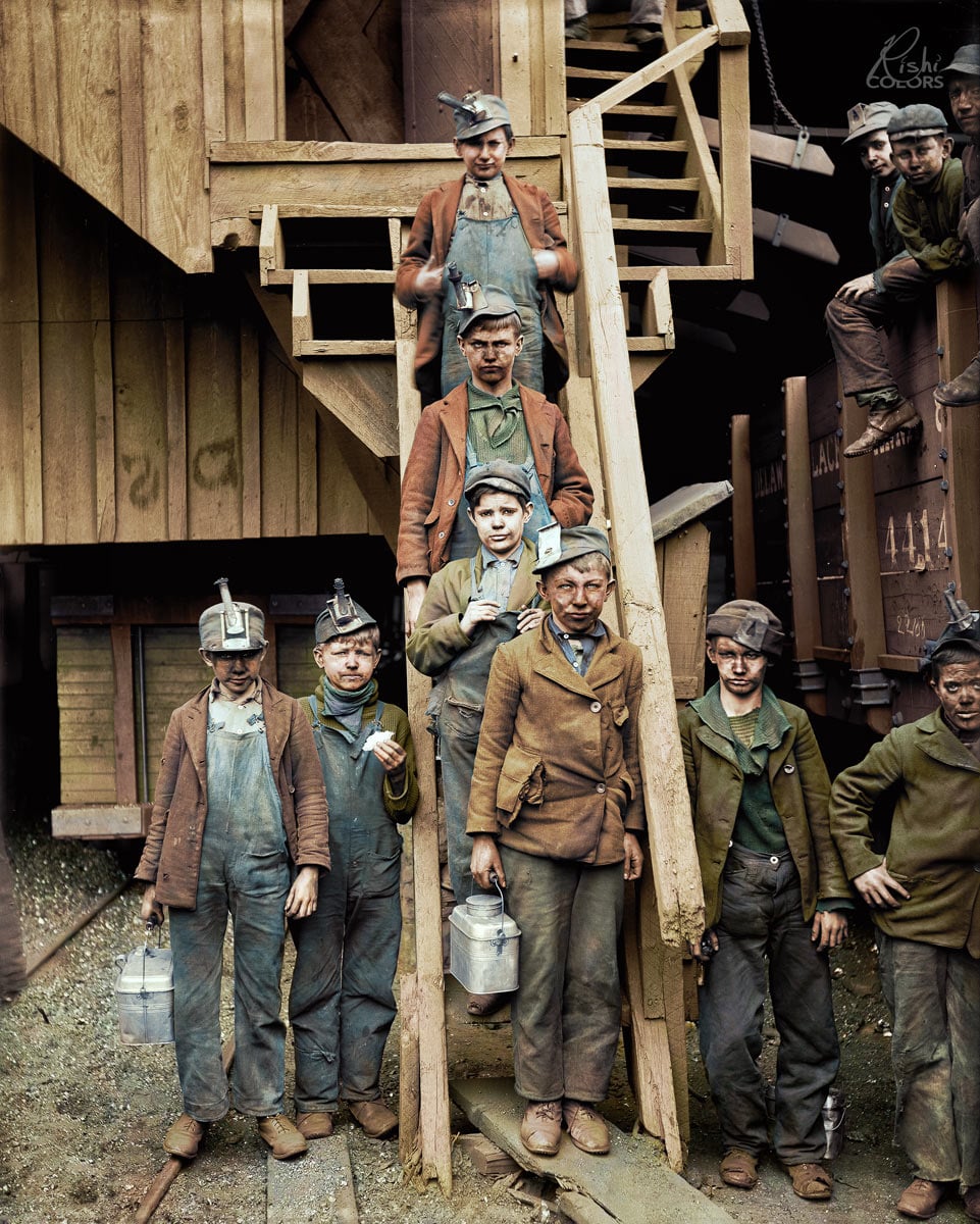 A group of young boys in work clothes and helmets stand and sit on wooden stairs outside an industrial building, holding lunch pails.