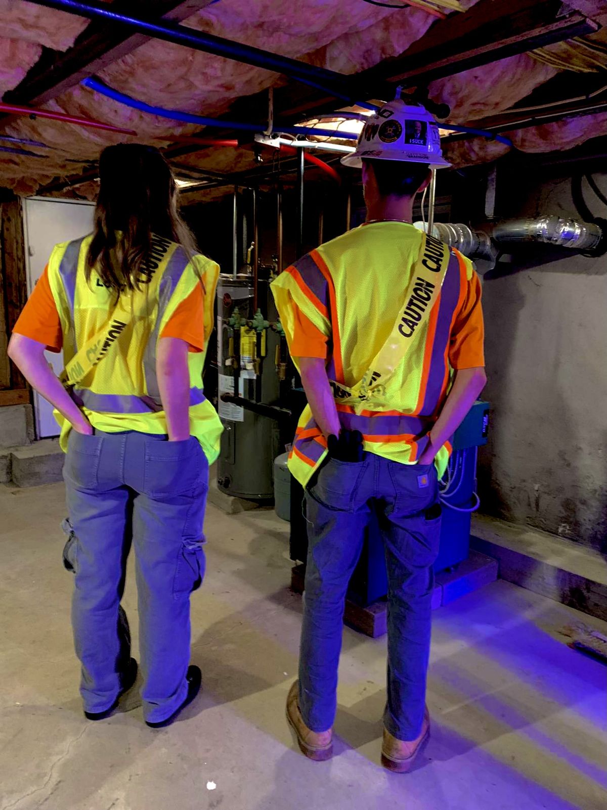 Two people wearing safety vests and helmets stand in a basement with exposed insulation, looking at machinery. Their hands are in their back pockets, and the area is lit with a blue-tinted light.