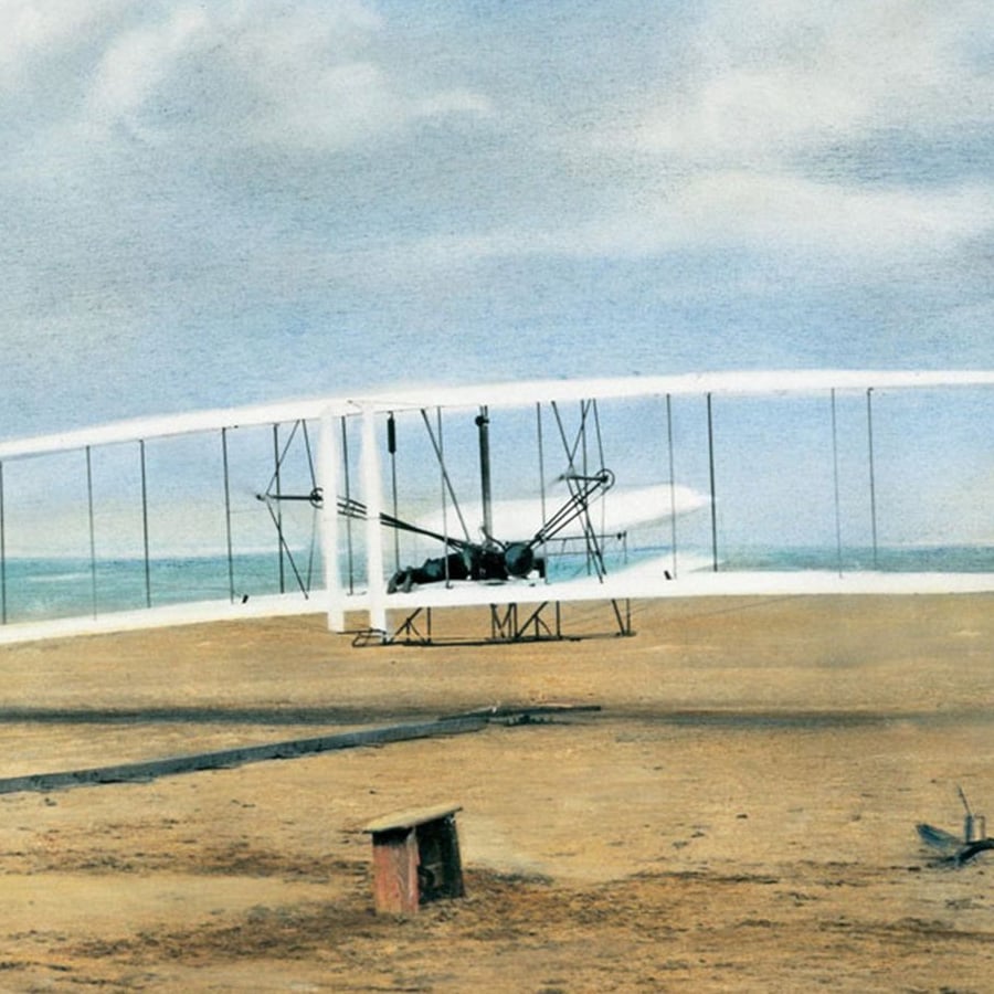 A vintage biplane with white wings stands on a sandy field under a partly cloudy sky, with the ocean visible in the background.