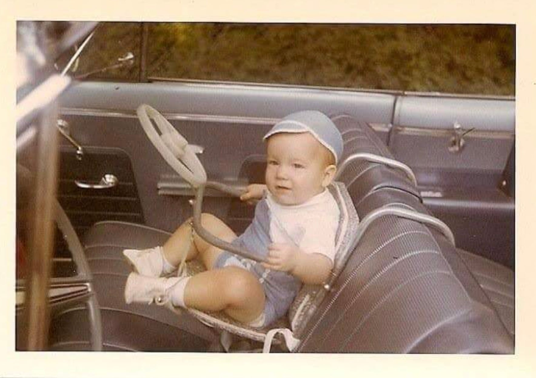 A baby wearing a cap sits in a car seat in the front seat of a vintage car, holding a toy steering wheel.