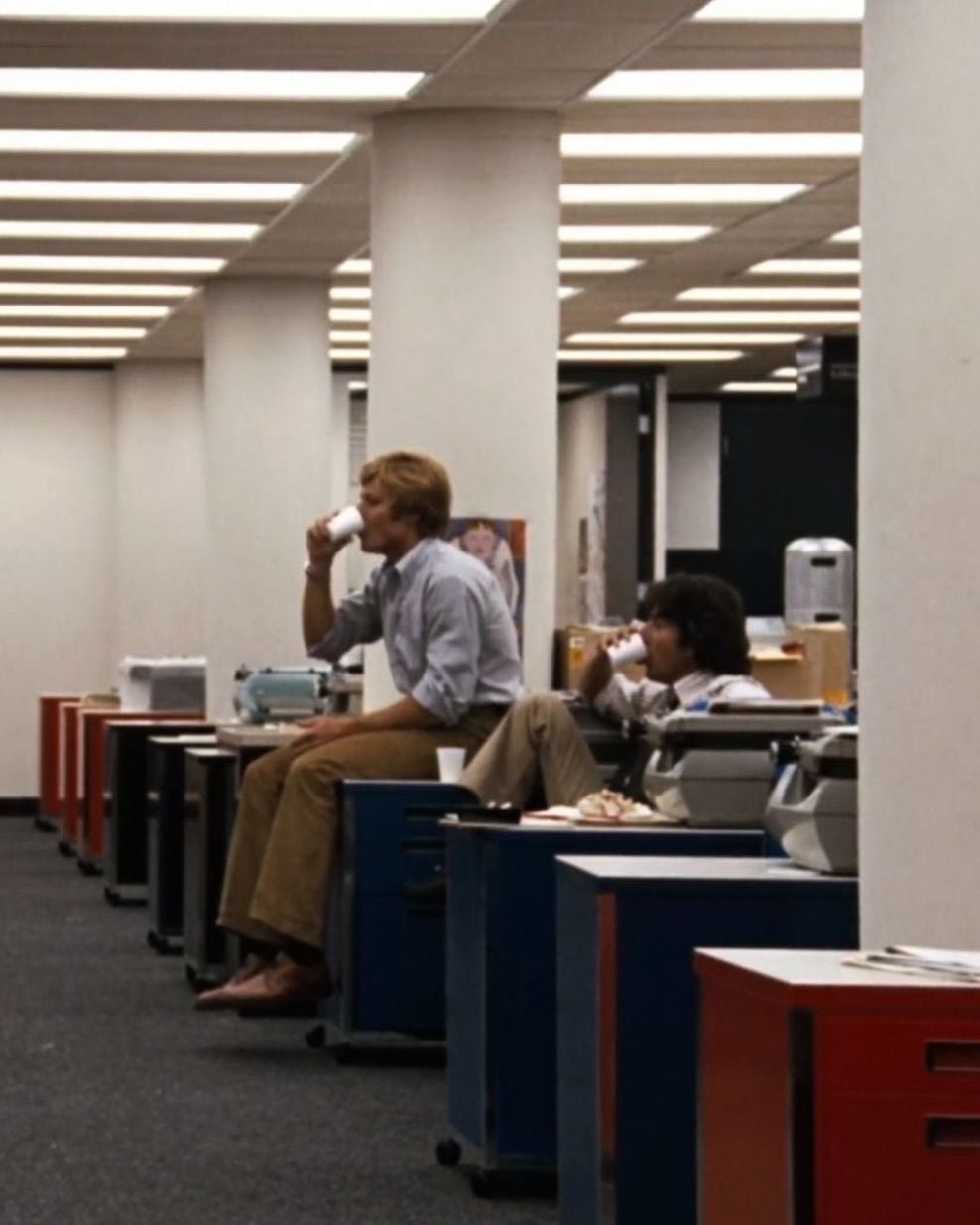 Two people sit on desks in an office, drinking from cups. The office has fluorescent lighting, white columns, and various office equipment and papers on the desks. The scene has a retro, 1970s atmosphere.