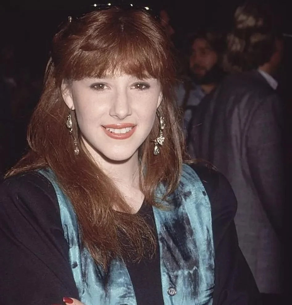 A woman with long red hair, wearing a blue patterned vest and dangling earrings, smiles at the camera. She stands in a dimly lit indoor setting with people blurred in the background.