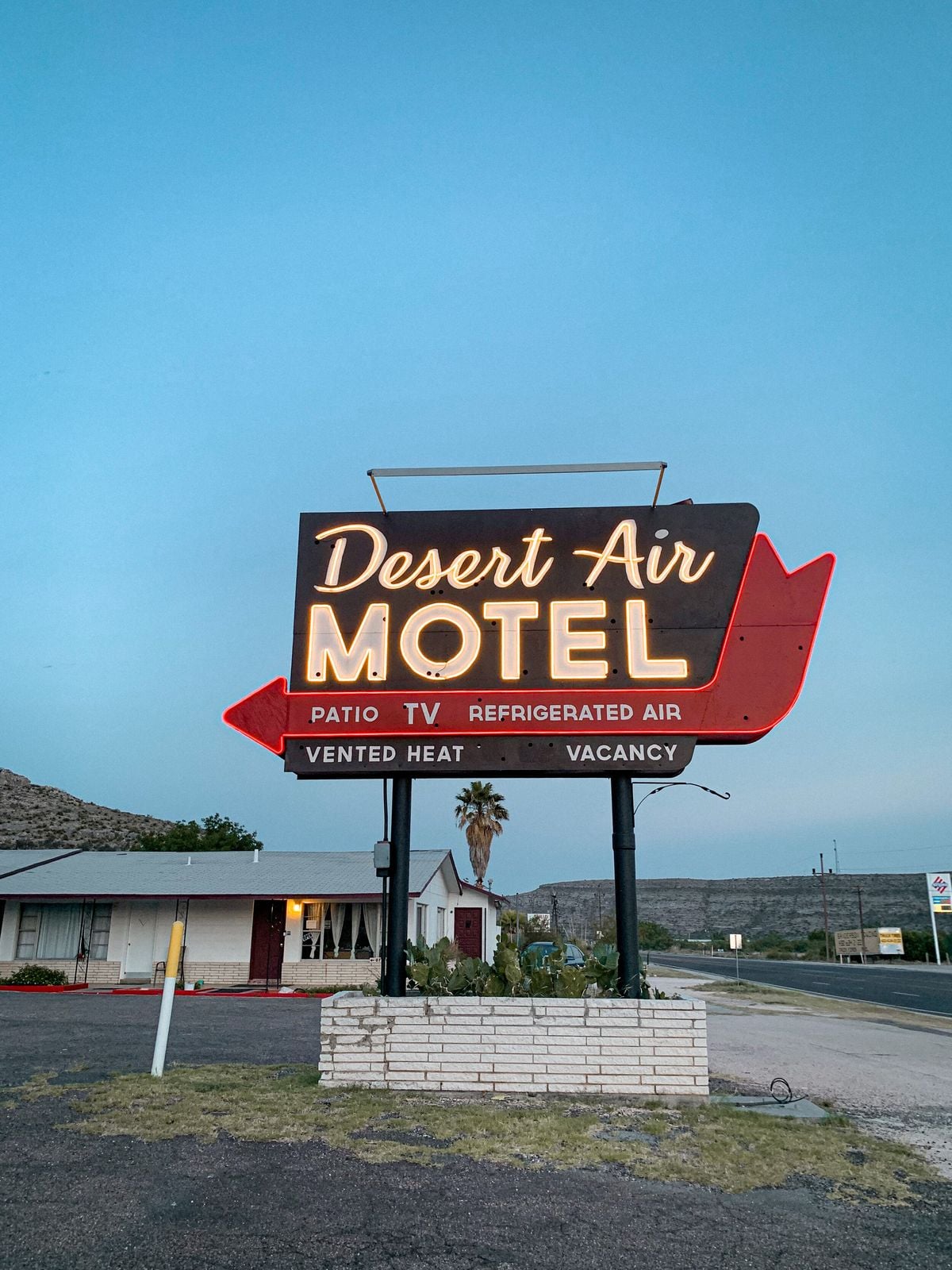 A vintage neon sign reads "Desert Air Motel" with "Patio TV Refrigerated Air" and arrows for "Vented Heat" and "Vacancy"; desert landscape and road in the background.