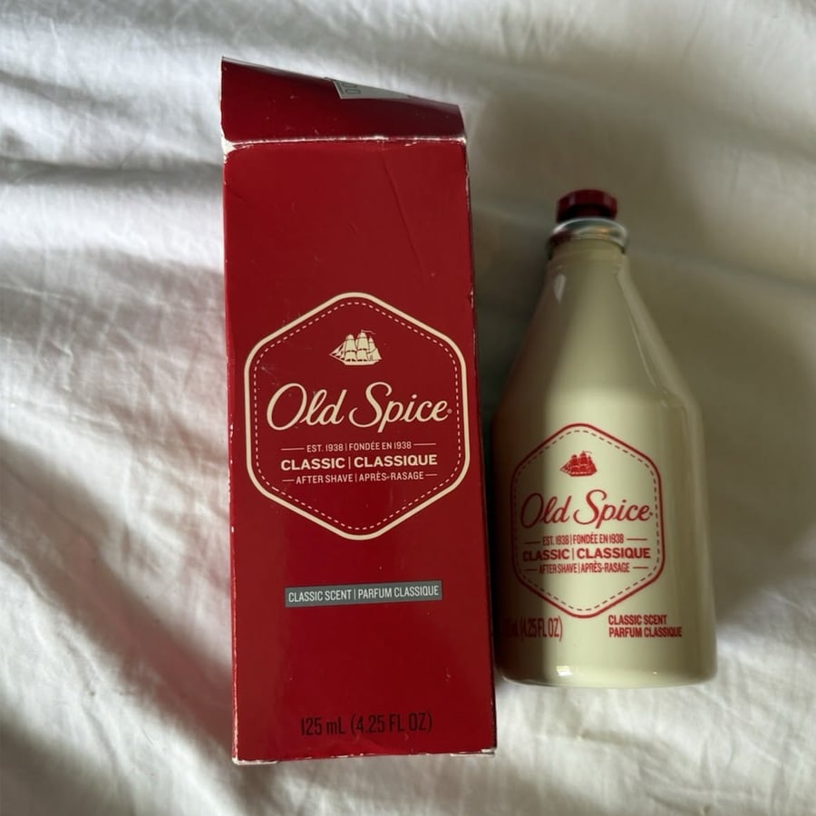 A red box and a matching vintage-style Old Spice after shave bottle sit on a white surface. The box and bottle both feature the Old Spice logo and classic scent labeling.