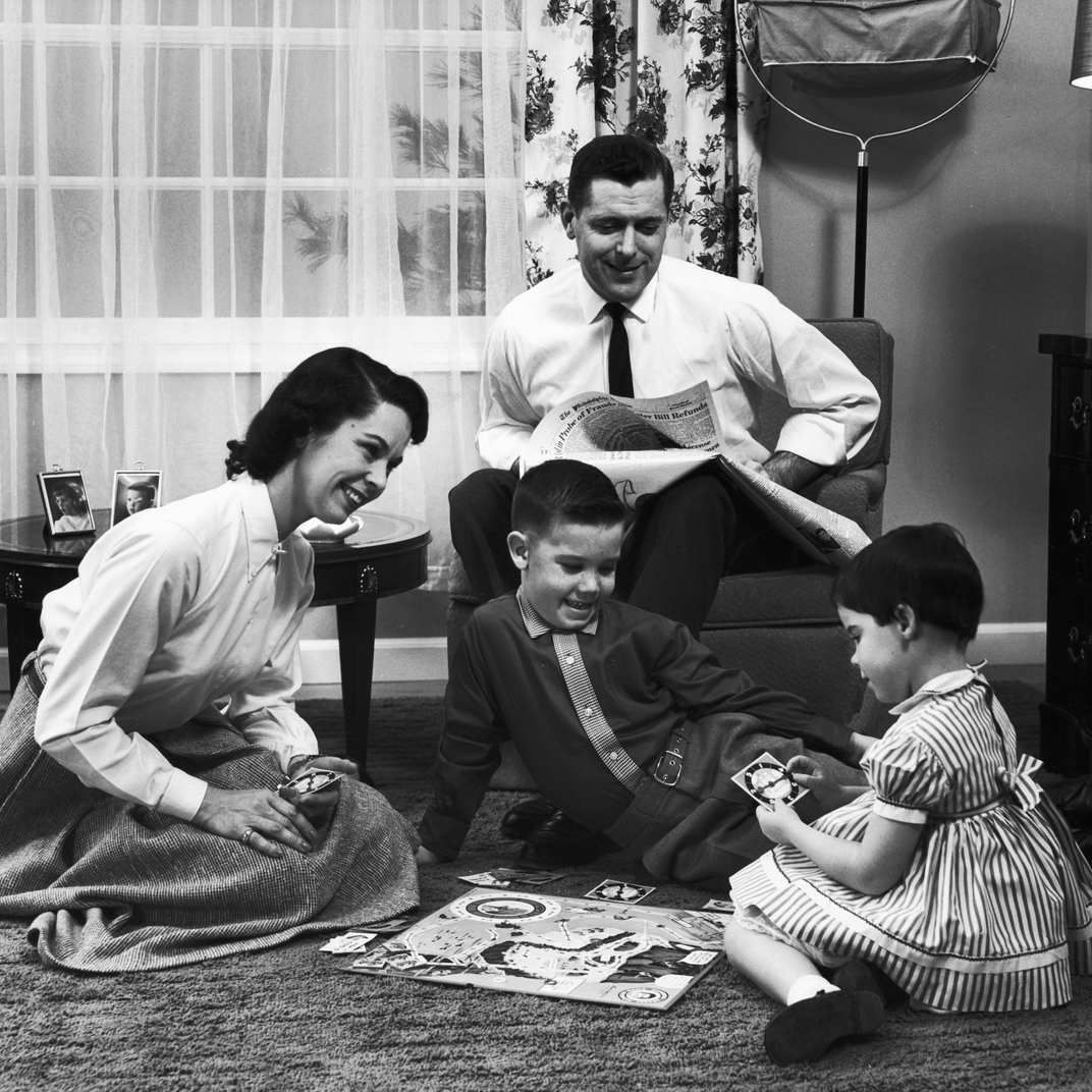 A smiling family of four plays a board game on the floor; the mother kneels, the father sits with a newspaper, and two young children laugh and hold game cards in a cozy living room.