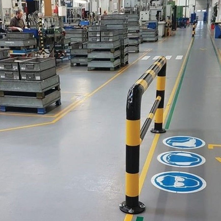Factory floor with safety barriers and stacked crates; marked pedestrian walkway with blue safety signs indicating helmets, gloves, and protective shoes required.