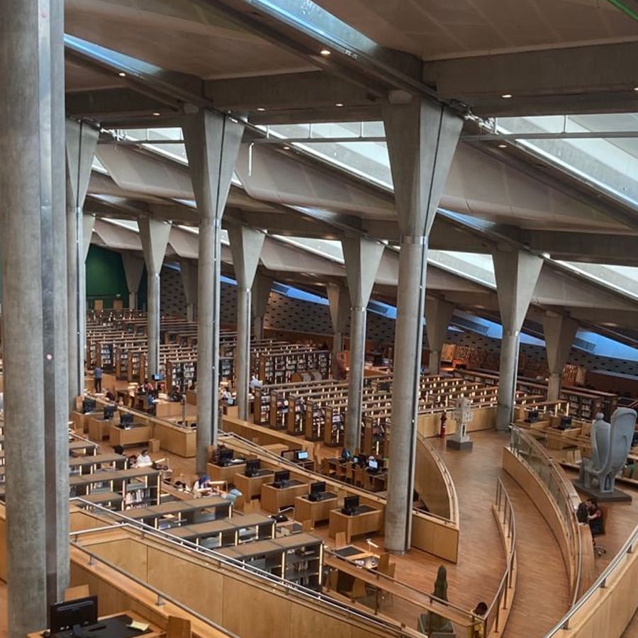 A spacious, modern library with high ceilings, large pillars, wooden desks, bookshelves, and computer stations. Natural light streams through angled windows, creating a bright, open atmosphere for studying and reading.