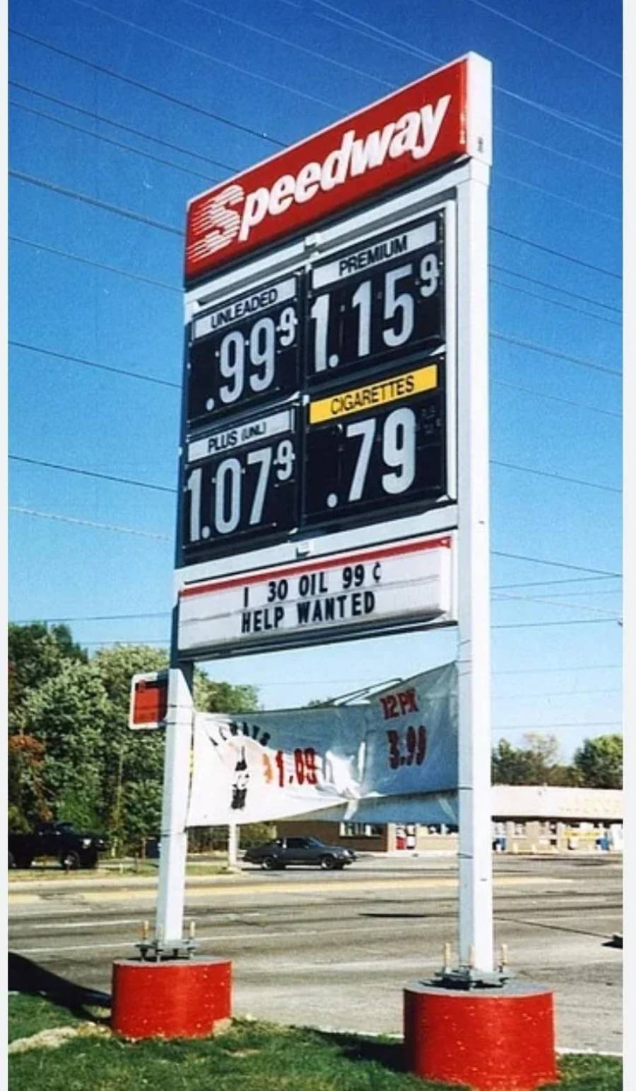 A Speedway gas station sign shows prices: Unleaded $0.99, Plus $1.07, Premium $1.15, Cigarettes $0.79. The sign also advertises “HELP WANTED” and “30 OIL 99¢.” Trees and a building are in the background.