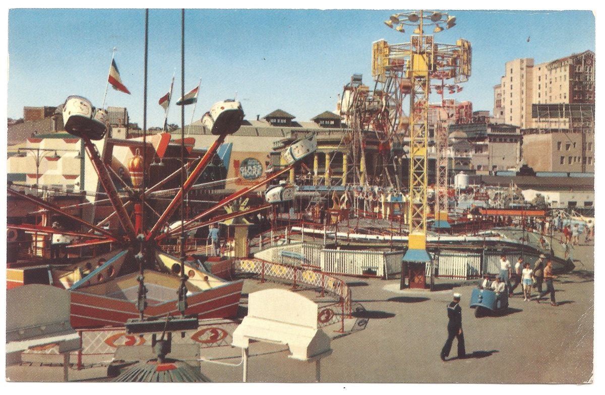 A vintage amusement park with colorful rides, including a Ferris wheel and spinning rides, is bustling with visitors. Buildings and flags are visible in the background under a clear blue sky.