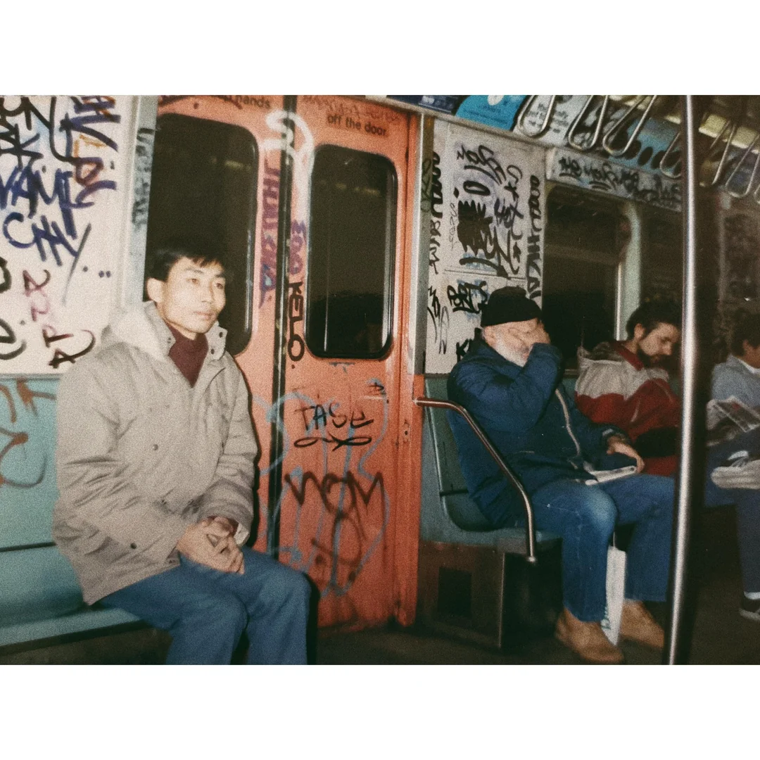 Three people sit on a graffiti-covered subway train; one looks ahead, another covers their face, and the third looks down, reading. The mood is subdued, and the subway car is heavily tagged with colorful graffiti.