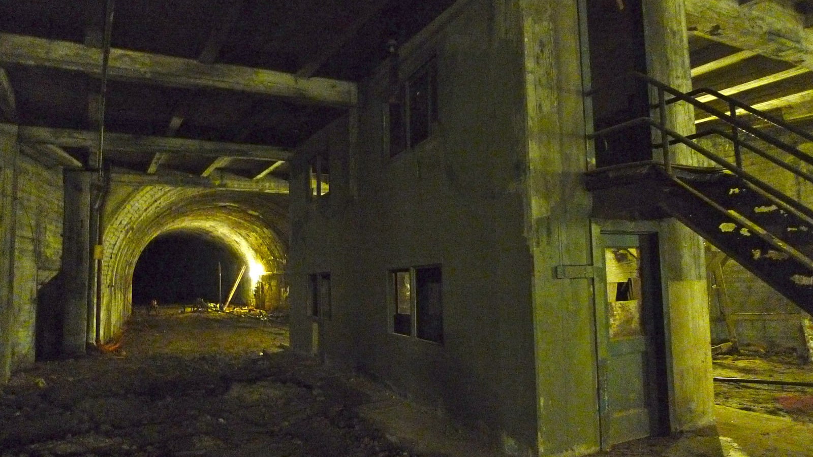 A dimly lit, abandoned underground tunnel with rough concrete walls and a small, two-story structure featuring a staircase and broken windows. Yellow lighting illuminates part of the tunnel entrance.