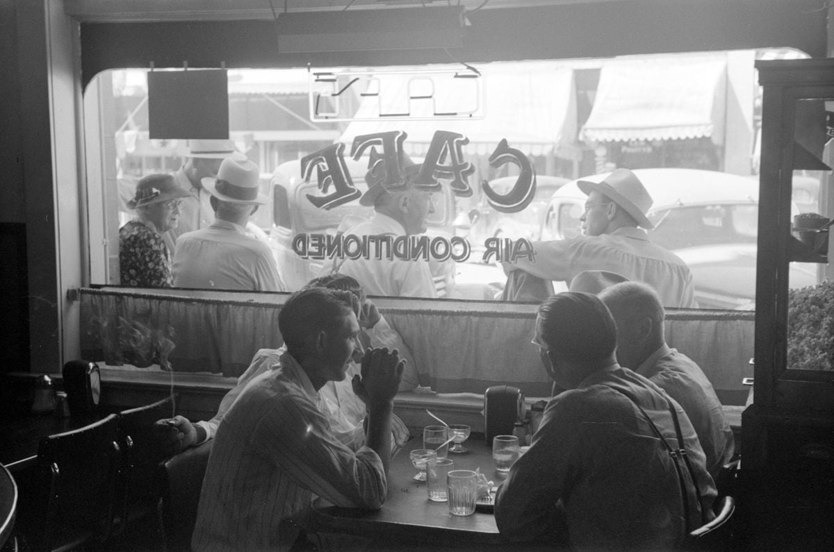 Black and white photo of men sitting at a café table, talking and drinking, with others visible outside through a window that reads “CAFÉ AIR CONDITIONED.” Vintage cars and storefronts appear in the background.