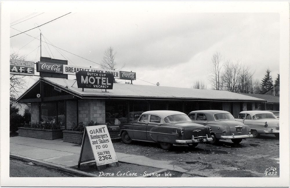 Black and white photo of Dutch Cup Cafe and Motel with vintage cars parked outside. A sign advertises “giant hamburgers and salads to go.” The diner has Coca-Cola signs and a "No Vacancy" motel sign.