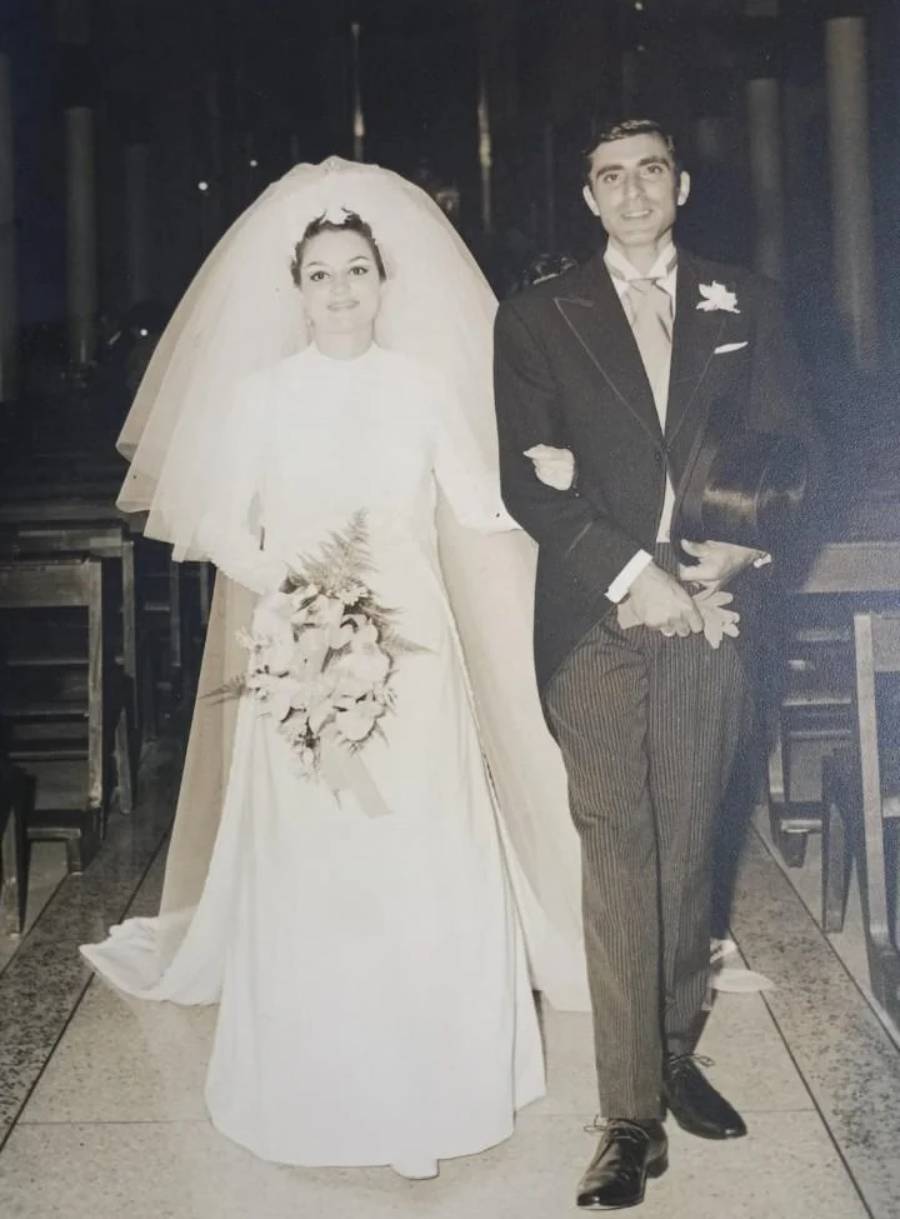 A vintage black-and-white photo of a bride in a long white wedding dress and veil holding a bouquet, arm-in-arm with a groom in a suit, walking down the aisle of a church.