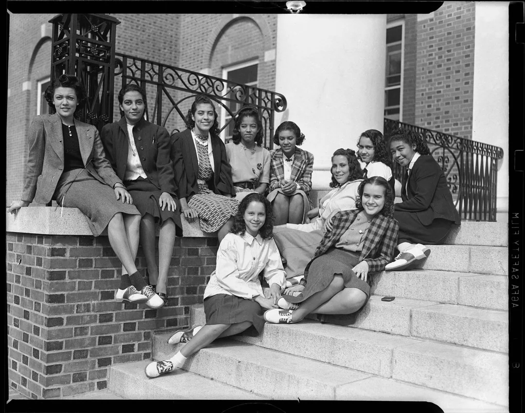 A group of ten young women sit and pose together on outdoor steps in front of a brick building, smiling and dressed in mid-20th-century clothing.