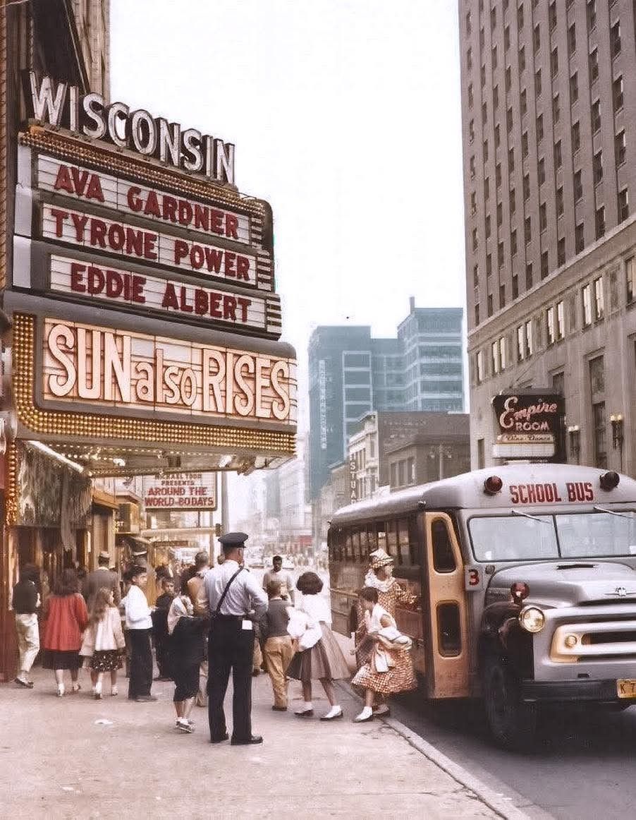 A vintage street scene shows children boarding a yellow school bus in front of the Wisconsin theater, whose marquee advertises “Sun Also Rises” starring Ava Gardner, Tyrone Power, and Eddie Albert. Pedestrians and shops line the sidewalk.