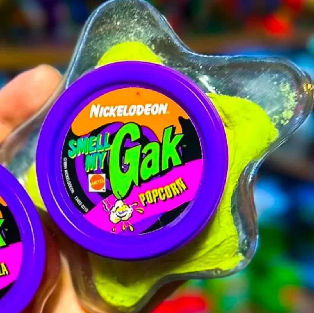 A hand holds a container of Nickelodeon Smell My Gak in a star-shaped package with a purple lid. The label reads "Popcorn" and the gak inside is bright yellow.