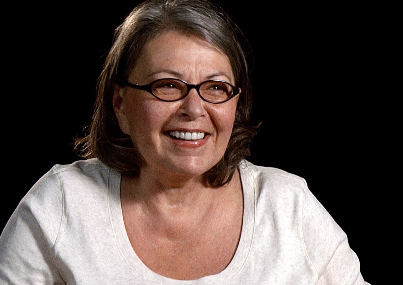 A woman with shoulder-length brown hair and glasses is smiling. She is wearing a light-colored top and is pictured against a plain black background.