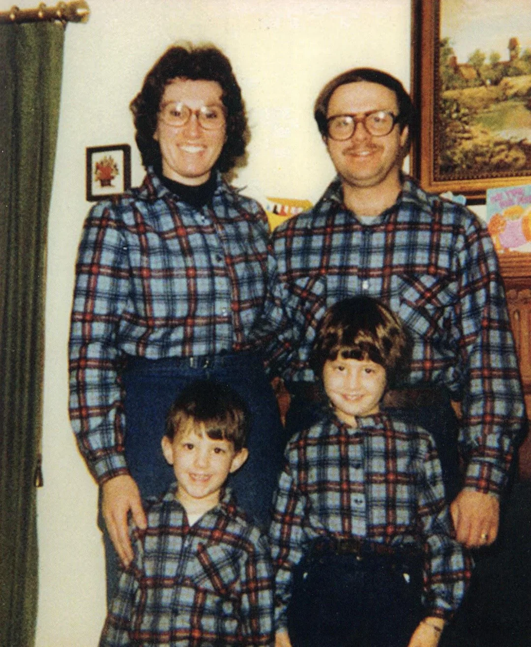 A family of four—two adults and two children—smile while wearing matching plaid shirts and glasses, standing indoors near a curtain with framed pictures and a painting on the wall behind them.