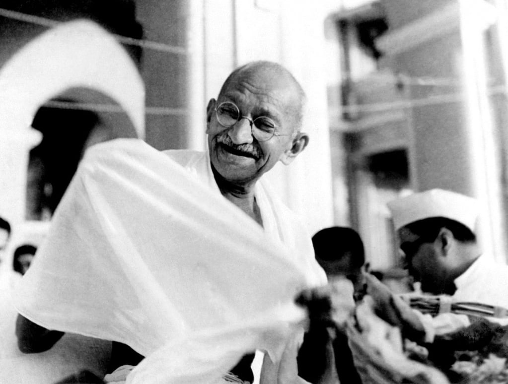 An elderly man with glasses and a mustache smiles while dressed in a simple white cloth. Other people are in the background, one wearing a white cap. The setting appears to be a public or outdoor gathering.