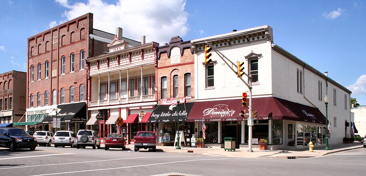 Historic downtown street with brick buildings, shops with awnings, parked cars, and a traffic light on a sunny day; blue sky with a few clouds above.
