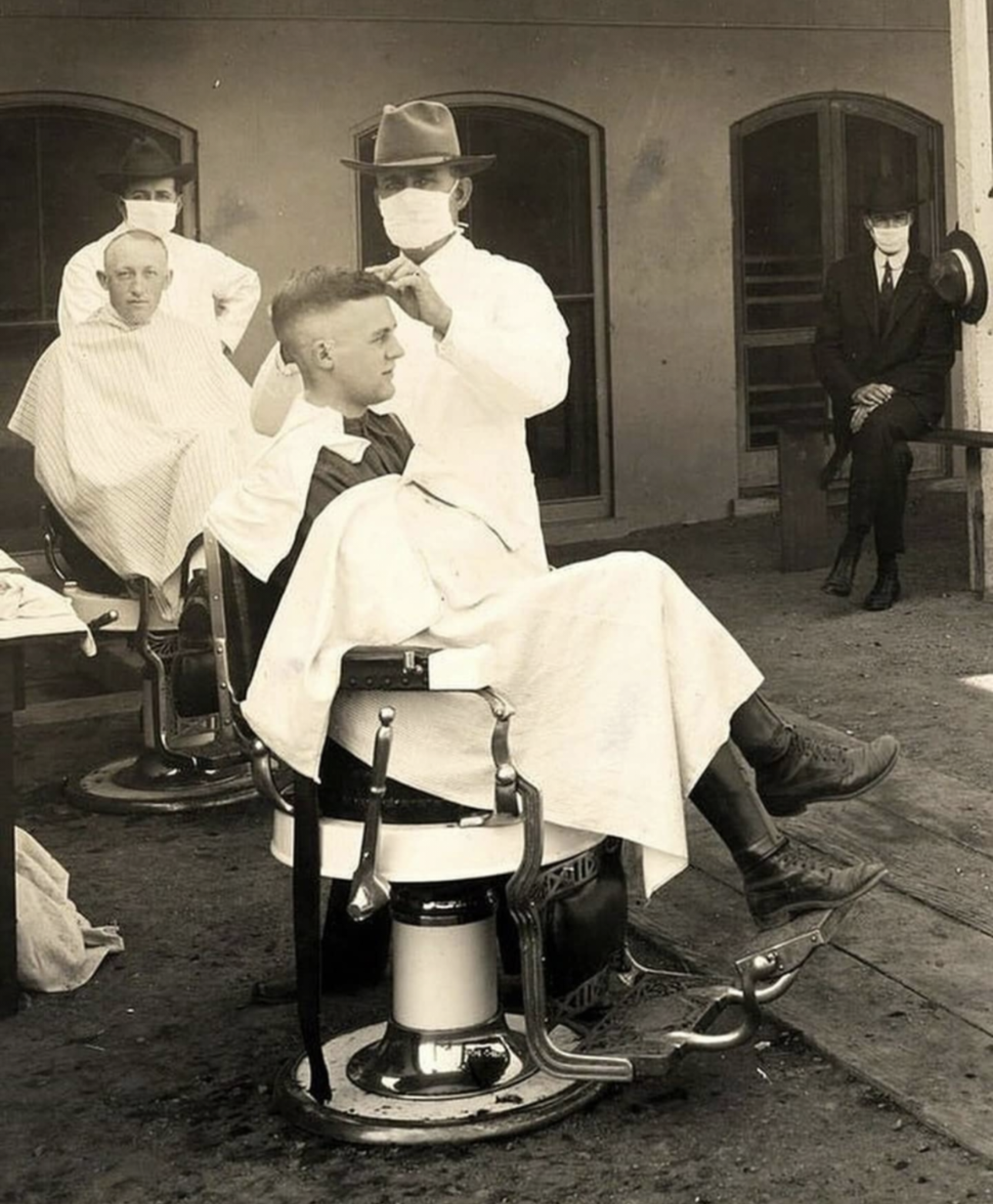 A barber wearing a mask cuts a man's hair at an outdoor barbershop, while another customer sits and a man in a suit waits nearby. The scene appears to be from the early 20th century.