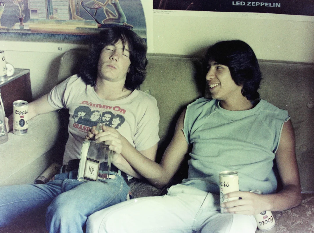 Two young men sit on a couch with drinks; one looks tired holding a bottle, the other smiles while holding his hand. Beer cans and band posters, including Led Zeppelin, are visible in the background.