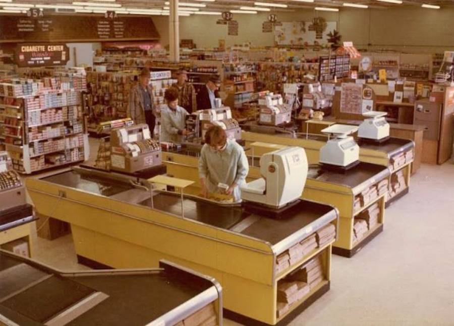 A vintage supermarket scene with yellow checkout counters, old-fashioned cash registers, and a few people shopping. Shelves stocked with groceries and a sign reading "Cigarette Center" are visible in the background.