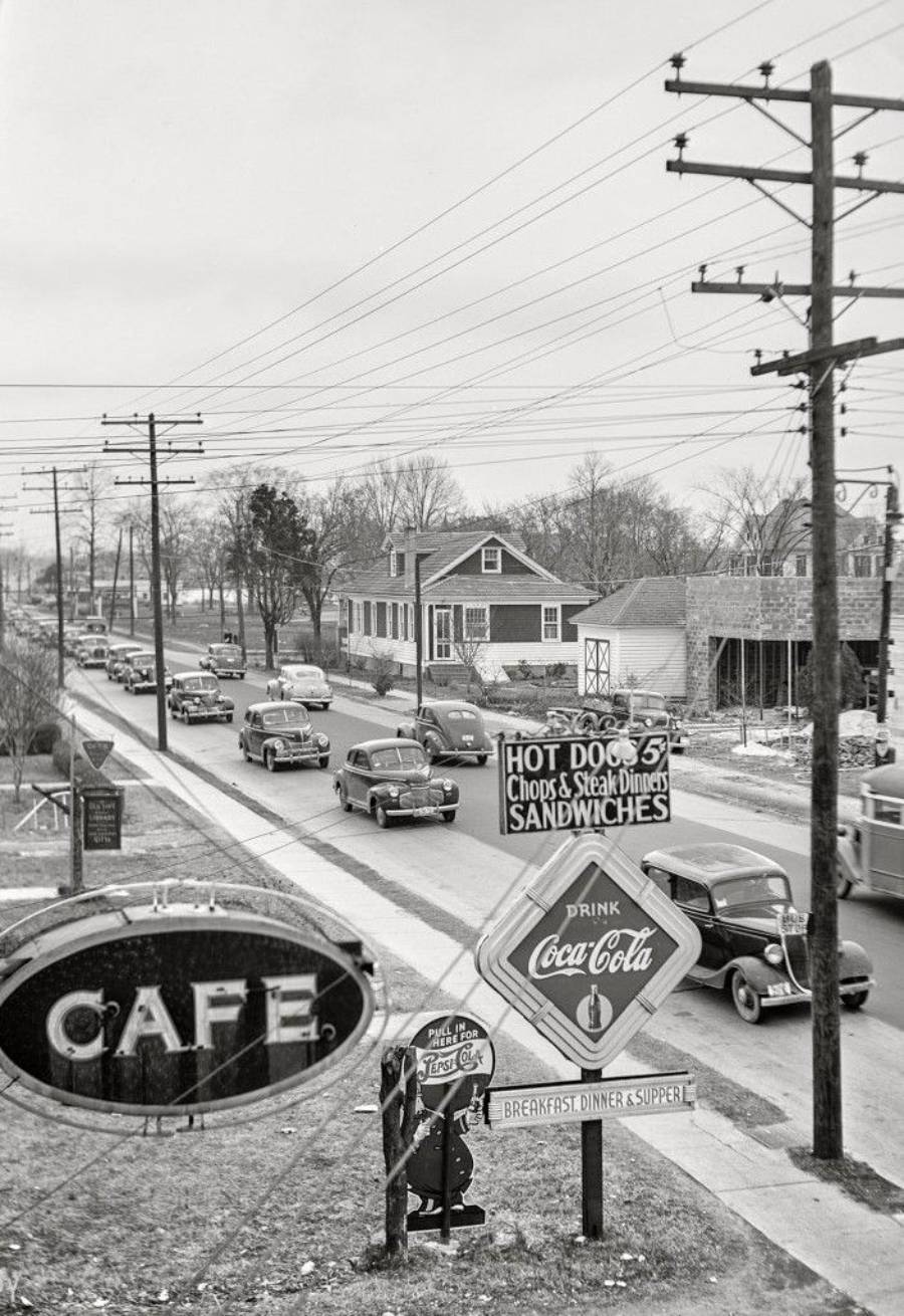 Black-and-white photo of a 1940s street scene with vintage cars, utility poles, a café, a Coca-Cola sign, and signs advertising hot dogs, steak dinners, sandwiches, and breakfast. Houses and trees line the street.