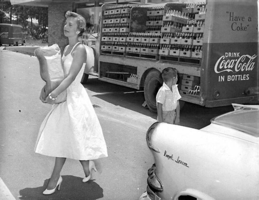 A woman in a white dress and high heels carries a grocery bag while walking past a young boy looking up, with a Coca-Cola delivery truck and a parked car labeled "Impala" in the background.