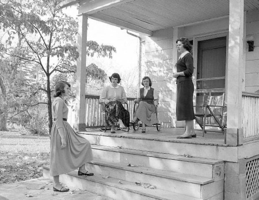 Four women are gathered on a wooden porch. Three sit on chairs, while one stands on the steps facing them. Trees and grass are visible in the background, and the scene has a relaxed, casual atmosphere.