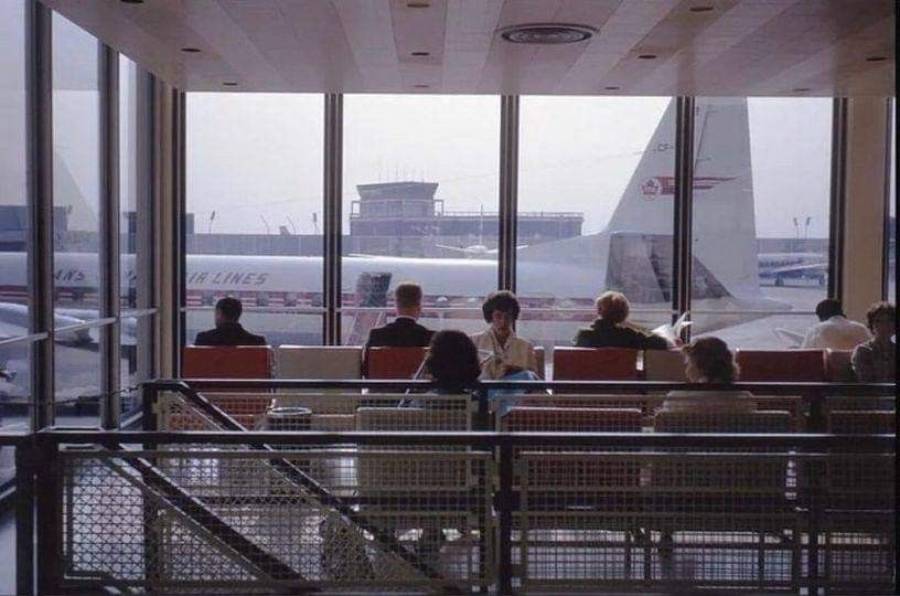 People sit in red chairs at an airport terminal, looking out large windows at airplanes parked outside. The scene appears calm, with natural light filling the waiting area.