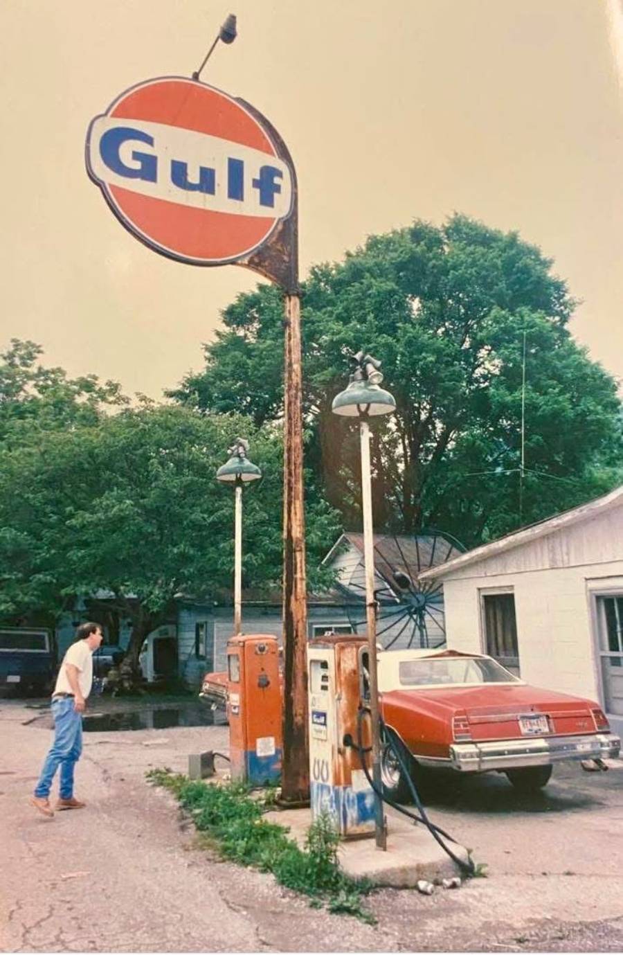 A man stands near old orange Gulf gas pumps beside a red vintage car parked in front of a white building, with large trees in the background.