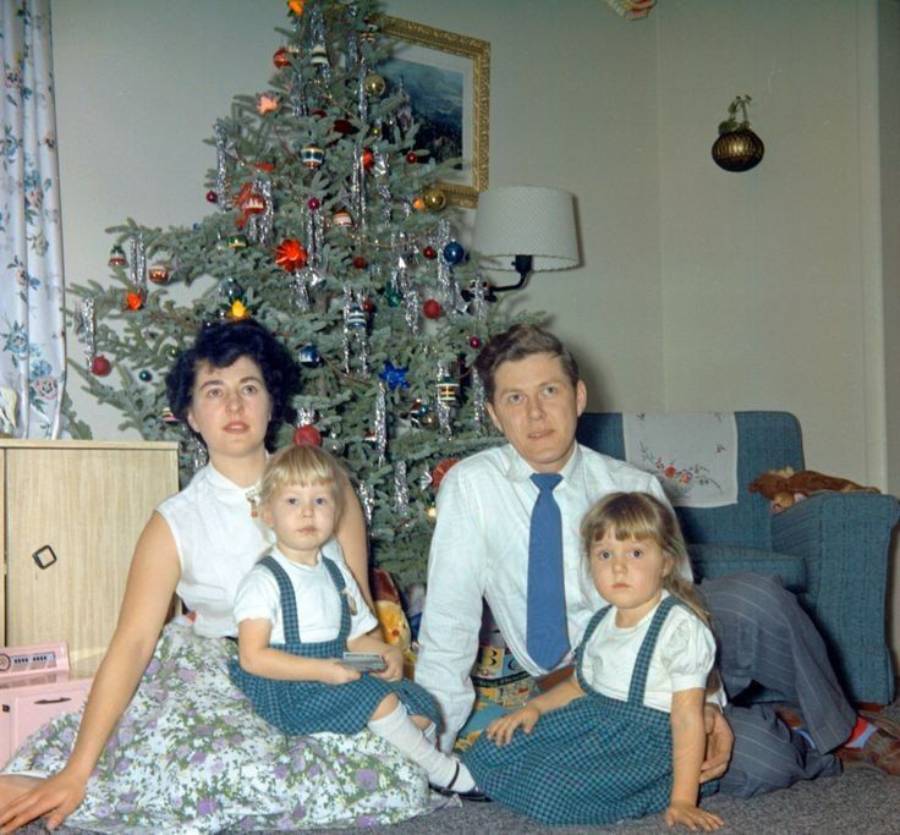 A family of four poses in front of a decorated Christmas tree. The parents sit with two young girls in matching blue plaid outfits. Festive ornaments decorate the tree and the room has a cozy, vintage feel.