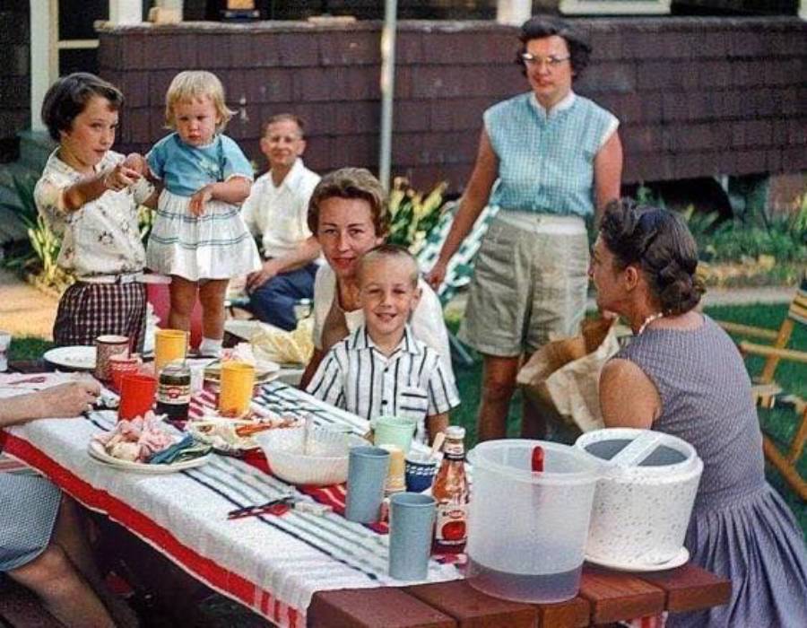 A group of adults and children gather at a picnic table outdoors, enjoying food and drinks. The table is covered with a red-and-white cloth and various dishes. Everyone appears relaxed and happy.